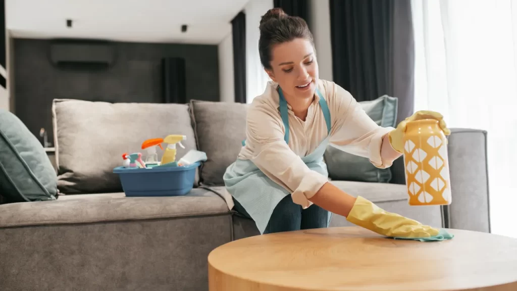 Mulher usando luvas de Limpeza do Ambiente e avental azul limpando uma mesa de centro em uma sala de estar, com produtos de limpeza sobre o sofá ao fundo.