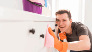 Homem sorridente limpando uma gaveta branca com pano rosa, usando luvas laranjas e segurando um borrifador. Ao fundo, balde roxo e acessórios de limpeza sobre um móvel branco, em um ambiente bem iluminado.