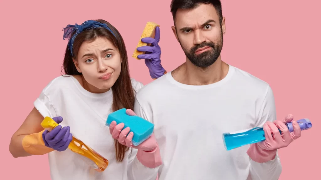Casal confuso segurando produtos e esponjas de limpeza, com expressão de dúvida, ilustrando o tema "Mitos e Verdades sobre Limpeza", em frente a um fundo rosa.